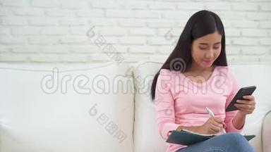 快乐的年轻亚洲女人在家学习，在手机上看短信或短信，微笑着坐在沙发上。